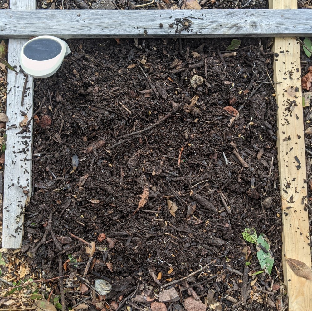A square foot garden square with prepared soil and a purple stone marker.