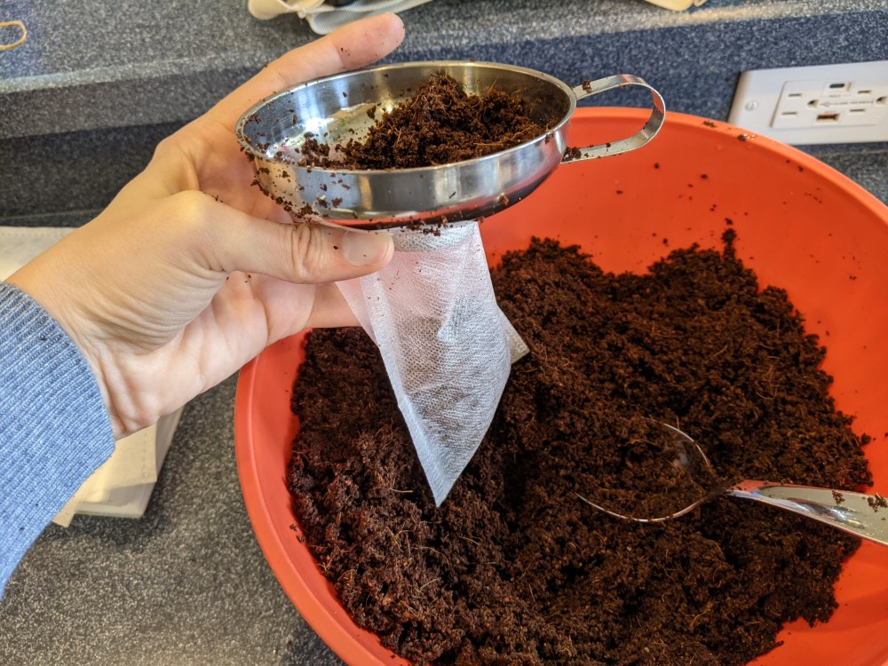 A bowl of seed starting mix and a spoon with a hand holding a canning funnel over a grow bag to fill the bag with soil.