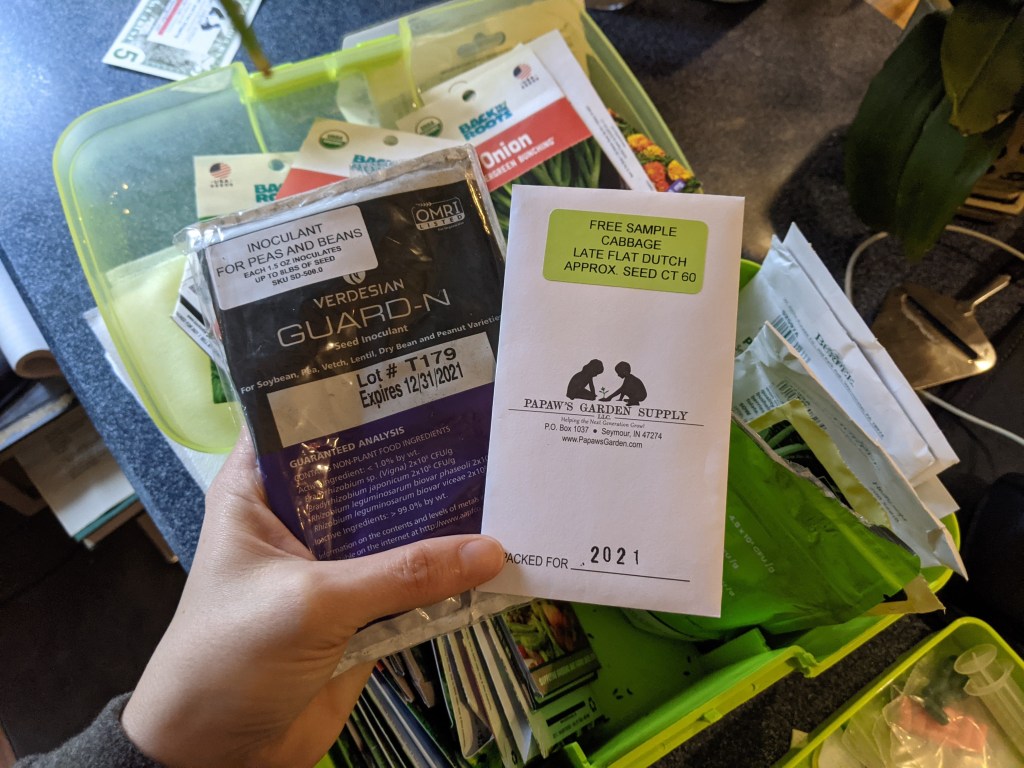 A hand holding a packet of soil inoculant for peas and beans and a packet of cabbage seeds. A box with seed packets in the background.