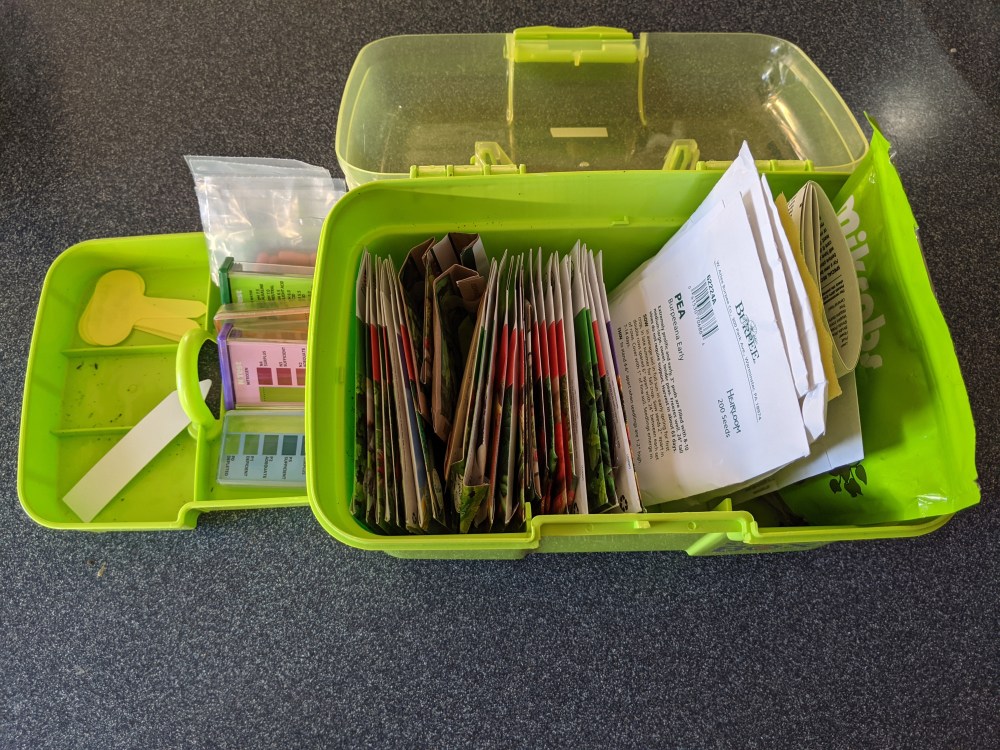 Small toolbox with lid open and tray to one side. Seed packets, plant labels, soil tests, and seeds can be seen inside.